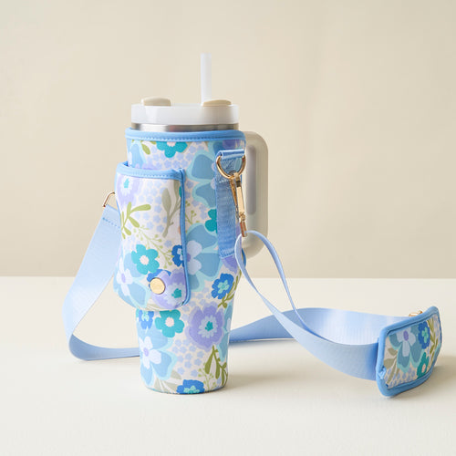 A large cup with a lid and straw sits in The Darling Effect’s Beyond Blooms Blue Green Tumbler Sling, a blue floral-patterned neoprene holder with matching adjustable strap, set on a light surface against a neutral background.