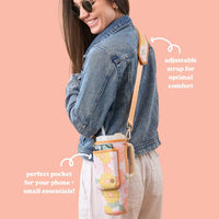 A smiling woman in sunglasses, a denim jacket, and light pants carries The Darling Effect’s Daisy Craze Peach Tumbler Sling—a floral neoprene sling bag with an adjustable strap, phone pocket, and versatile features.