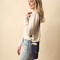 A smiling woman in a cream sweater and jeans carries The Darling Effect's Pretty Posies Navy Tumbler Sling, featuring a floral design, phone pocket, and adjustable strap.