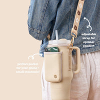 A person in a denim jacket and light pants carries The Darling Effect’s Tan Tumbler Sling, a neoprene tumbler carrier with an adjustable strap, beige color, phone side pocket, and text emphasizing comfort and utility.