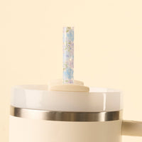 A close-up of the-darling-effect’s Secret Garden Blue 40 oz Tumbler Straw Set, a white tumbler with metal rim and BPA-free reusable clear straw featuring a pastel floral pattern, shown against a pale beige background.