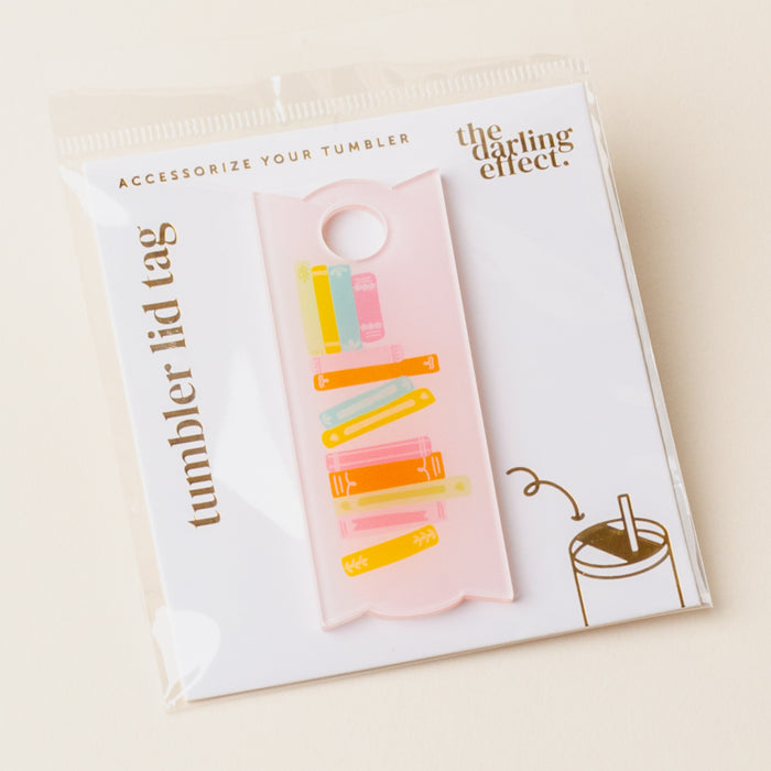 A pink, translucent acrylic Books Stack Tumbler Lid Tag shaped like a long rectangle with a hole at the top, printed with a stack of colorful books, inside clear packaging.
