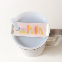 A rectangular Books Stack Tumbler Lid Tag featuring an illustration of stacked colorful books, attached to a clear straw on a white tumbler.