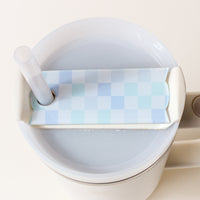 A tumbler filled with liquid, topped with The Darling Effect’s Check Peach Tumbler Lid Tag—a checkered blue and white lid featuring a hole for a clear straw and a convenient side handle that enhances both style and functionality.