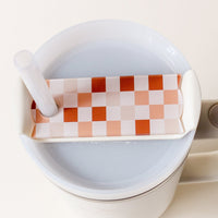 A close-up of a white cup with a light drink, topped with The Darling Effect’s Check Brown Tumbler Lid Tag, which has an orange-and-white checkered pattern and a hole for a clear straw. The cup has a right-side handle and resembles a personalized tumbler.