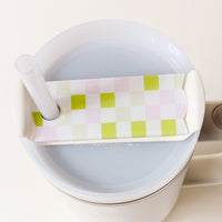 A white cup with a light gray drink has a Check Green Tumbler Lid Tag from The Darling Effect—a checkered pink, green, and white lid with a straw hole—ideal for personalizing your Stanley Quencher 2.0.