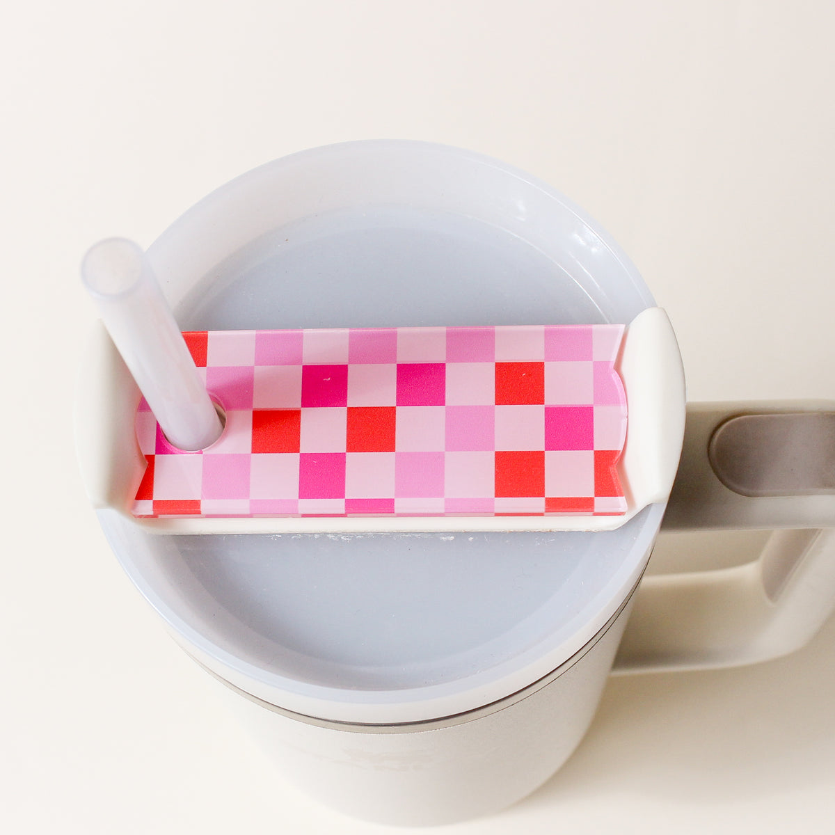 A large white cup with a handle, topped with a pink and red checkered lid featuring a hole for a clear straw, sits on a light surface. The Hot Pink Checker Tumbler Lid Tag by The Darling Effect adds an extra touch of charm, making this piece perfect for fans of the brand.