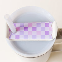 A cup with a white handle features The Darling Effect’s Check Purple Tumbler Lid Tag, a pastel purple and pink checkered Stanley Quencher 2.0 lid with a straw hole and inserted straw, topping off a light-colored drink.