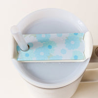 A clear plastic straw emerges from a white travel mug lid, decorated with The Darling Effect’s Daisy Craze Aqua Tumbler Lid Tag—ideal for fans of personalized accessories or pairing with Stanley Quencher 2.0 lids.