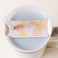 The Darling Effect’s Daisy Craze Peach Tumbler Lid Tag, featuring pastel pink, yellow, and orange flowers, is clipped to a Stanley Quencher lid with a straw on a white mug—a charming accessory for your tumbler.