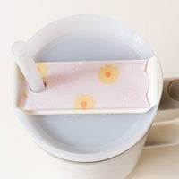 A close-up shows a large white cup with a light drink, topped with The Darling Effect's Darling Daisy Peach Tumbler Lid Tag—a pink floral-patterned straw cover that keeps the straw in place. The background is plain off-white.