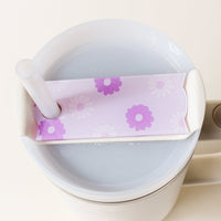 A white tumbler from The Darling Effect is topped with a rectangular Darling Daisy Purple Tumbler Lid Tag featuring a pink and purple floral design, complete with a straw through the lid’s hole for added style.