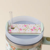 The Darling Effect’s Floral Haven Pink Tumbler Lid Tag, featuring a floral pattern and a hole for a clear straw, sits atop a pastel personalized tumbler—both decorated with matching floral designs.
