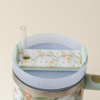 A close-up of a The Darling Effect tumbler with a transparent straw and the Floral Haven Sage Tumbler Lid Tag, featuring green leaves and small white and orange flowers on a light background.