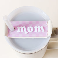 A white tumbler with a handle and straw features The Darling Effect's Mom Tumbler Lid Tag—a pink checkered tag that reads "mom" in white letters—attached to its Stanley Quencher 2.0 lid, filled with a light-colored drink.