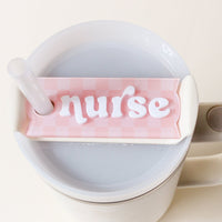 A white straw fits through The Darling Effect's Nurse Tumbler Lid Tag in pale pink checkered acrylic with white puffy "nurse" letters, made for Stanley Quencher 2.0 lids—cute accessory for travel mugs.