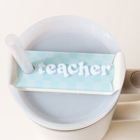 The Darling Effect’s Teacher Tumbler Lid Tag is a light blue and white checkered cover with “teacher” in raised white letters—a charming accessory to top your mug or tumbler.