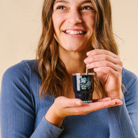A smiling woman in a blue top cheerfully holds and stirs the-darling-effect's Garden Blooms Navy 2 oz Tiny Tumbler filled with a dark drink.