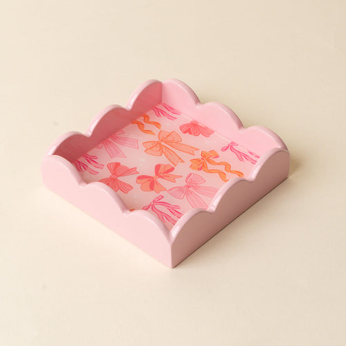 The Blushing Bows Scallop Trinket Tray by the-darling-effect is a square, light pink tray with scalloped edges and a playful pink and orange bow pattern, ideal for organizing jewelry or accessories.