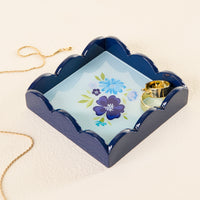 The Dainty Floral Navy Scallop Trinket Tray by the-darling-effect, a square, scallop-edged blue tray with a floral design, holds two gold rings while a gold necklace rests beside it on a cream surface.