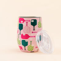 The Darling Effect’s Pickleball Party Wine Tumbler—a pink, insulated cup featuring vibrant paddle and ball designs—is pictured with its spill-resistant lid beside it on a light beige background.