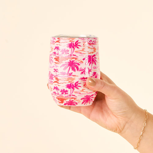 A manicured hand holds The Darling Effect's Paradise Palms Wine Tumbler, featuring a pink and orange palm tree pattern on beige, with triple-wall insulation and a spill-resistant lid.