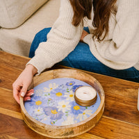A person in a cream sweater and blue jeans places the-darling-effect's Bouquet Beauty Purple Large Round Wood Enamel Tray with a lit candle on a wooden table.