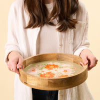A woman in a white shirt holds the-darling-effect's Bouquet Beauty Sage Large Round Wood Enamel Tray, filled with water and floating orange and white flowers, softly lit against a neutral background.
