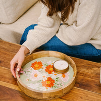 A woman in a cream sweater and blue jeans sits on a couch, reaching for the-darling-effect Bouquet Beauty Sage Large Round Wood Enamel Tray with a floral design beside a stack of white coasters on the wooden table.