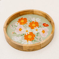 The-darling-effect’s Bouquet Beauty Sage Large Round Wood Enamel Tray features orange, white, and pink floral designs on a light green background, displayed on a white surface.