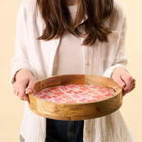 A person in a white shirt and dark jeans holds the-darling-effect Blushing Bows Large Round Wood Enamel Tray with a pink and orange leaf-patterned liner. The person's face is not visible.