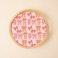 The Blushing Bows Large Round Wood Enamel Tray by the-darling-effect features a pink surface with red and pink bows, small white stars, and a plain light beige background.