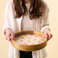 A person in a white shirt holds the-darling-effect's Endless Daydream Cream Large Round Wood Enamel Tray, which features a floral base with small pink and orange flowers on a light background. The person's face is not shown.