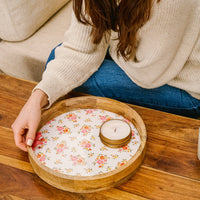 A person with long brown hair in a cream sweater and blue pants holds the-darling-effect's Endless Daydream Cream Large Round Wood Enamel Tray with a floral pattern and candle, seated at a wooden table by a light-colored couch.
