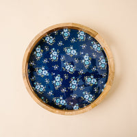 The Endless Daydream Navy Large Round Wood Enamel Tray by the-darling-effect features a dark blue surface with light blue flowers and green leaves, shown on a beige background.