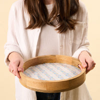 Someone holds the-darling-effect's Petal Parade Blue Large Round Wood Enamel Tray, showcasing its delicate floral pattern, against a plain background.