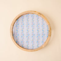 The-darling-effect’s Petal Parade Blue Large Round Wood Enamel Tray features blue flowers and green leaves on a white and light blue striped background, beautifully displayed on a beige surface.