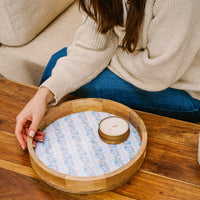A person in a cream sweater and blue jeans sits on a couch, reaching for the-darling-effect’s Petal Parade Blue Large Round Wood Enamel Tray with a floral pattern and coasters on a wooden coffee table.