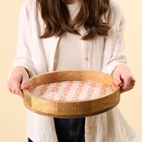 A person in a white shirt holds the-darling-effect Petal Parade Pink Large Round Wood Enamel Tray, featuring a pink floral and striped pattern, against a light beige background.