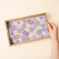 A hand holds the-darling-effect's Bouquet Beauty Purple Rectangle Wood Enamel Tray, showcasing a floral design with purple, white, and yellow blooms on a lavender background, set on a light cream surface.