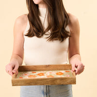A person in a white top and denim jeans holds the-darling-effect’s Bouquet Beauty Sage Rectangle Wood Enamel Tray, featuring a light green background with a vibrant orange and pink floral pattern.