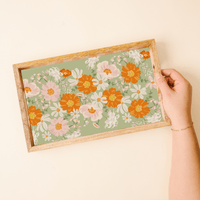 The-darling-effect’s Bouquet Beauty Sage Rectangle Wood Enamel Tray showcases a retro floral pattern in orange, pink, and white on a sage green background, elegantly displayed in-hand for decorative organization.