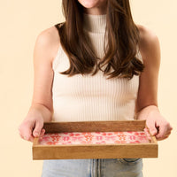 A woman in a sleeveless white top and jeans holds the-darling-effect's Blushing Bows Rectangle Wood Enamel Tray, featuring a pink and red floral design. Her face isn't shown, with a plain light beige background behind her.