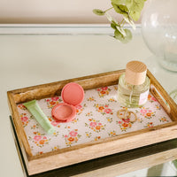 The-darling-effect’s Endless Daydream Cream Rectangle Wood Enamel Tray holds a green tube, an open pink compact, a glass perfume bottle with wooden cap, and two gold rings on a glass surface next to a vase of green leaves.