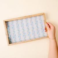 A hand holds the-darling-effect's Petal Parade Blue Rectangle Wood Enamel Tray, featuring a blue and white floral pattern with a wooden base that accentuates its delicate design.