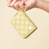 A hand holds The Darling Effect’s Green Check Zip Around Wallet—compact and stylish with a green checkered pattern, a secure zipper closure, and a handy keyring. The wallet is adorned with a small metallic heart-shaped emblem in the corner, seamlessly blending elegance and functionality.