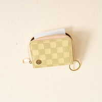 A compact, green checkered zip-around wallet from The Darling Effect, featuring a keyring and shown partially unzipped to reveal a white card inside, resting on a light-colored surface.