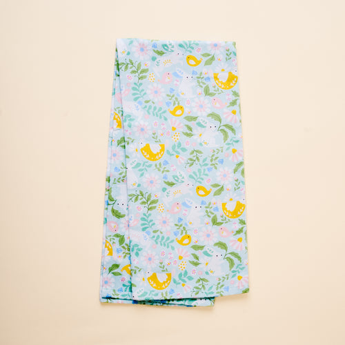 The Hoppy Days Tea Towel by The Darling Effect features light blue fabric adorned with yellow birds, green leaves, and pastel flowers, shown folded on a beige background.