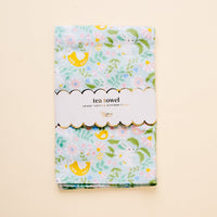 The Hoppy Days Tea Towel by the-darling-effect features a light blue floral and lemon design. It’s folded with a white scalloped label reading "tea towel" and "100% cotton kitchen towel," shown against a cream background.