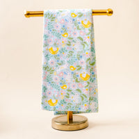 The Hoppy Days Tea Towel by the-darling-effect, featuring a vibrant pattern of yellow birds, flowers, and leaves on light blue fabric, is displayed neatly on a gold metal stand against a neutral background.